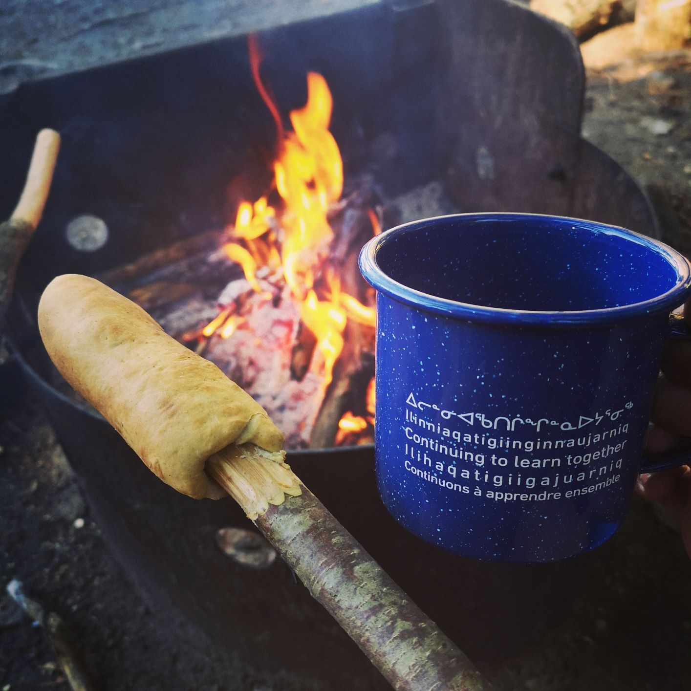 Bannock on a stick is shown beside a campfire and a blue mug that says continuing to learn together in several Indigenous languages